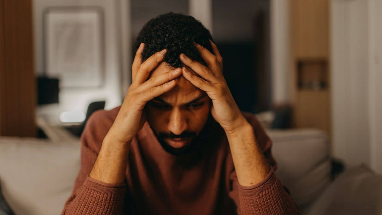 A man in a maroon sweater sits with his head held in both his hands.