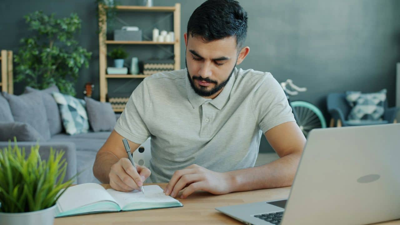 A man with a beard sits at a desk writing in a notebook near a laptop.