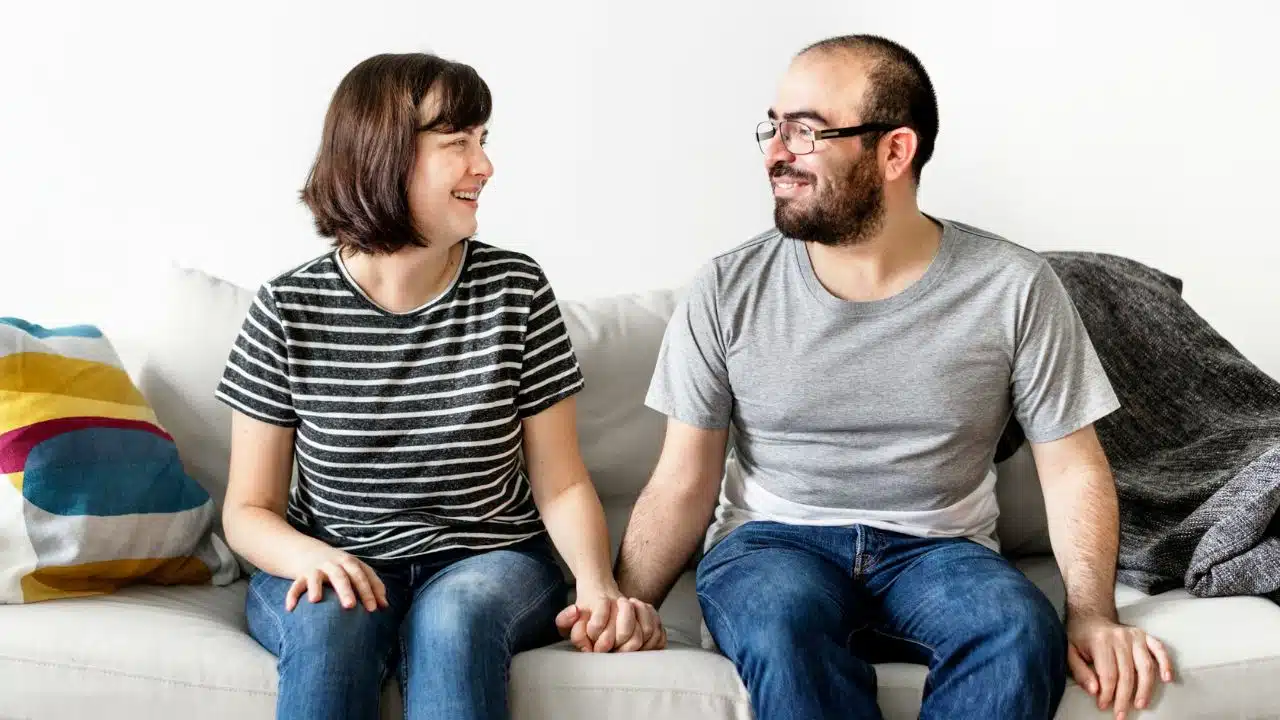 A man and woman sit on a couch, holding hands and smiling at each other.