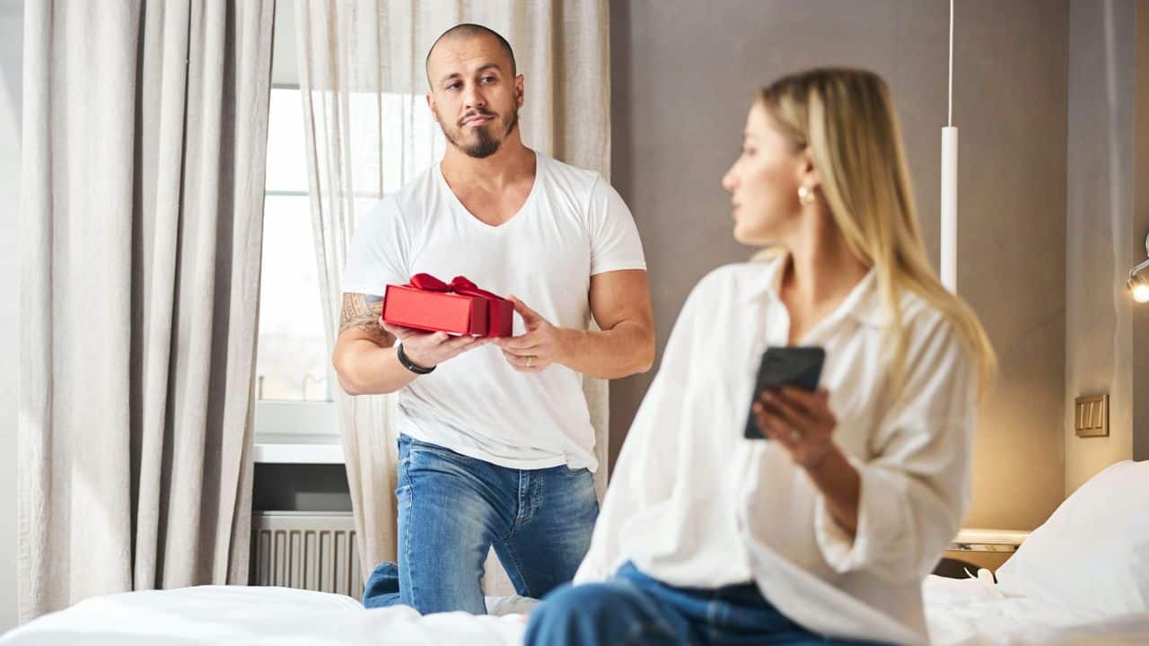 A man kneels with a red gift box while a woman looks at her phone.