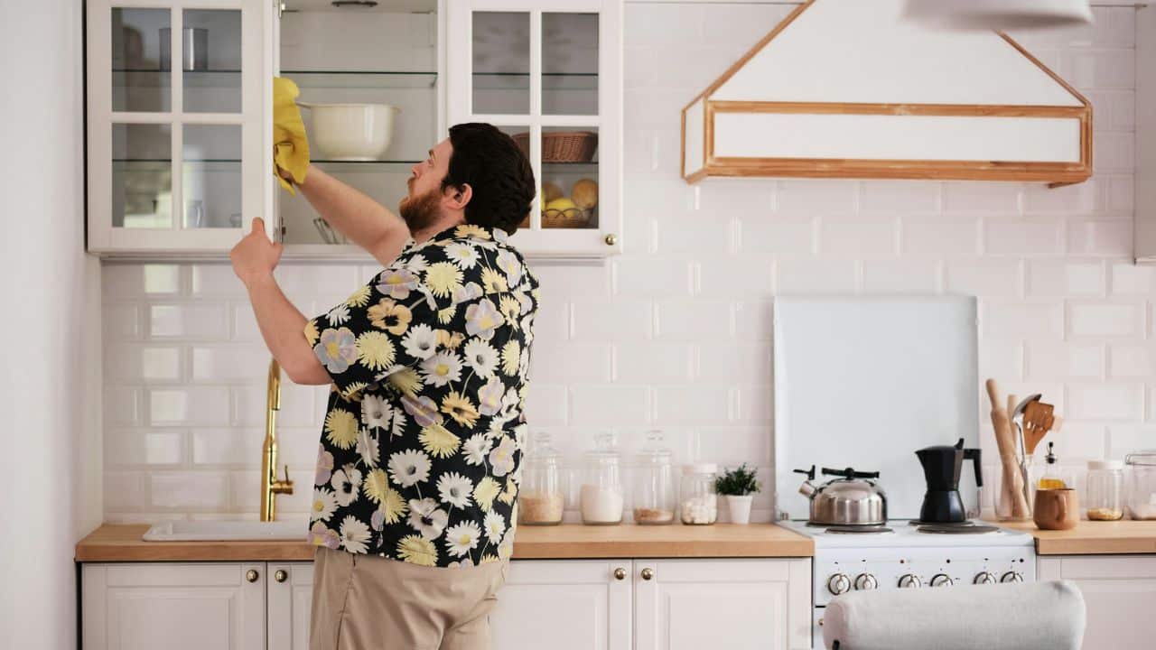A man in a floral shirt wipes a white kitchen cabinet with a yellow cloth.