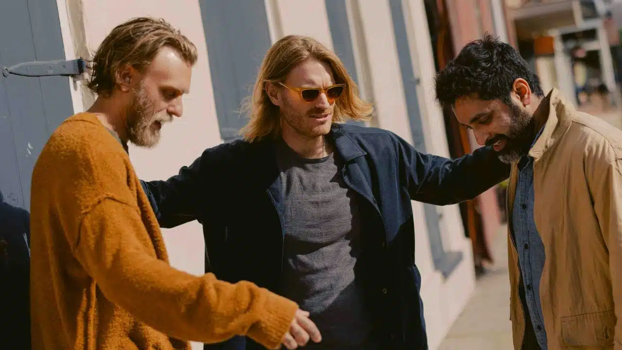 Three men stand together outdoors, with the center man resting his hands on their shoulders.