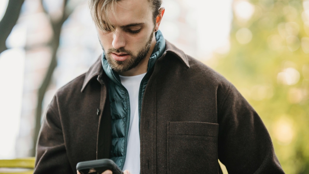 A man with a beard and brown jacket looks down at his phone outdoors.