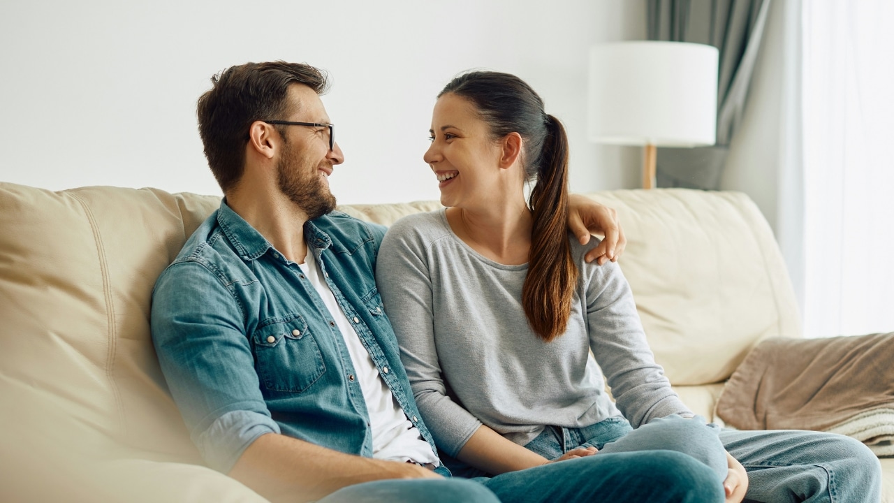 A man and woman sit on a beige sofa, smiling and looking at each other.