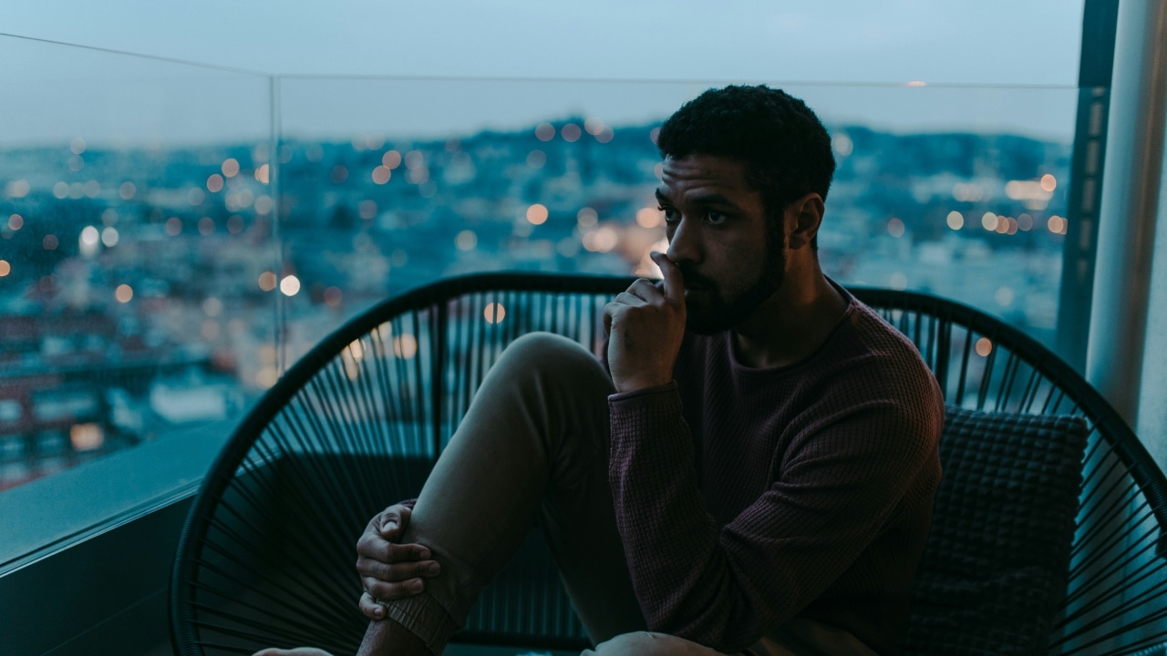 A man sits on a balcony chair at dusk overlooking a glowing city skyline.