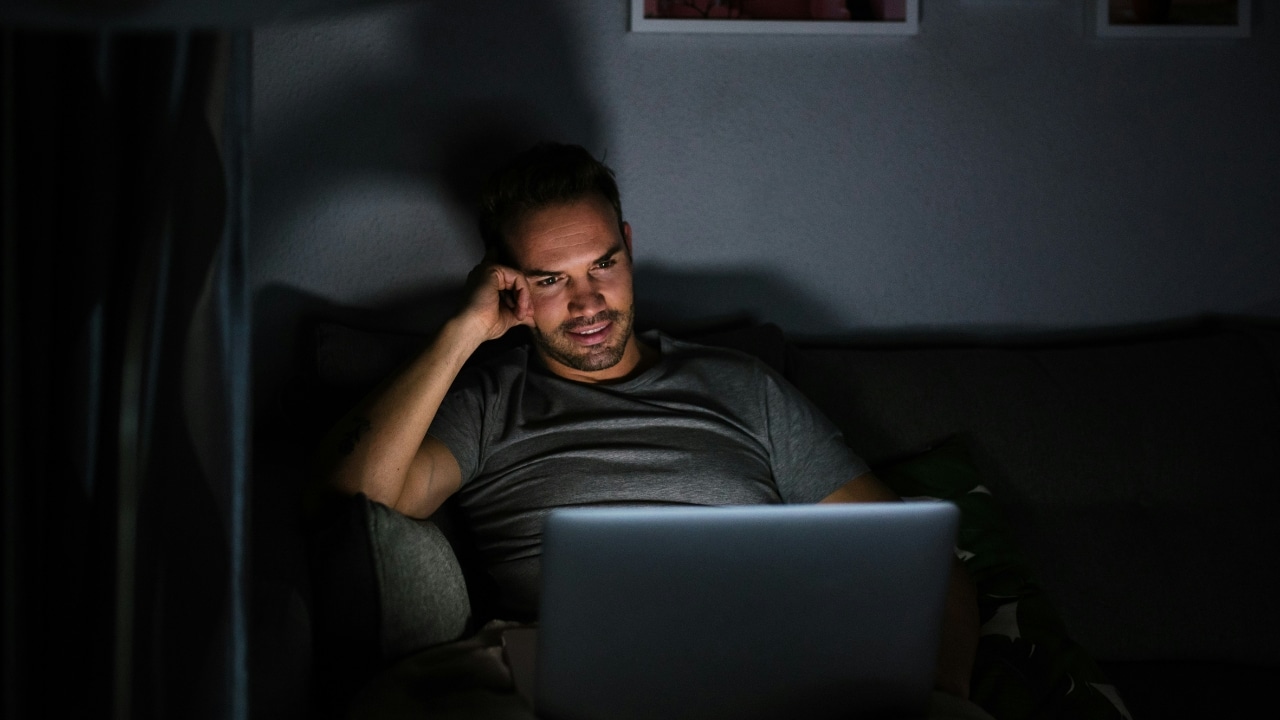 A man sits on a dark couch at night looking at a glowing laptop screen.