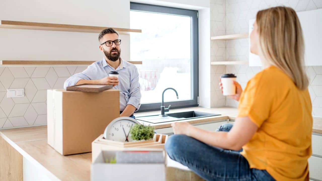 A bearded man in glasses holds a cup and talks to a woman in a kitchen.