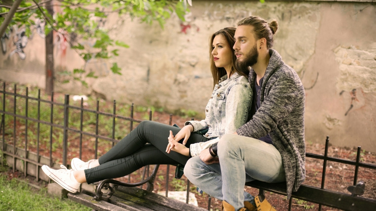 A man with a bun and a woman in a denim jacket sit on a bench.