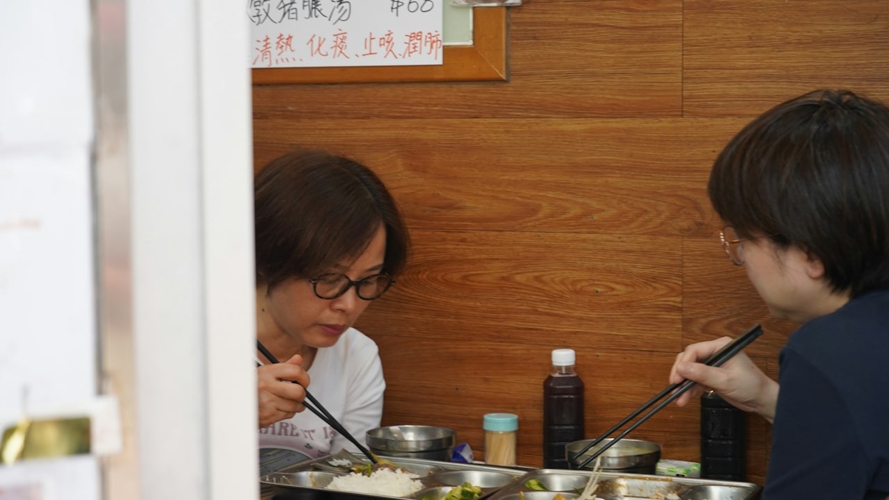 Two people eating with chopsticks out of metal trays at a simple food stall with a wooden wall and a menu sign with Chinese text.