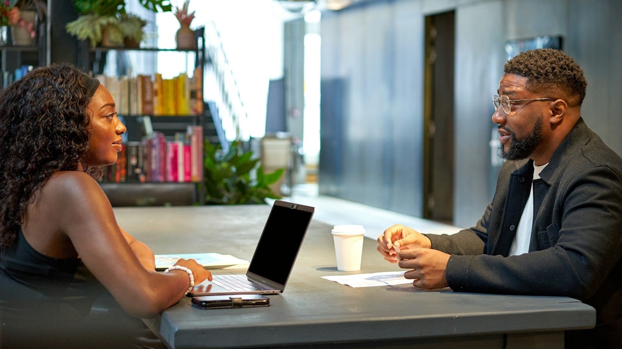 A woman and man sitting across a table from each other, engaged in a discussion in a modern office.