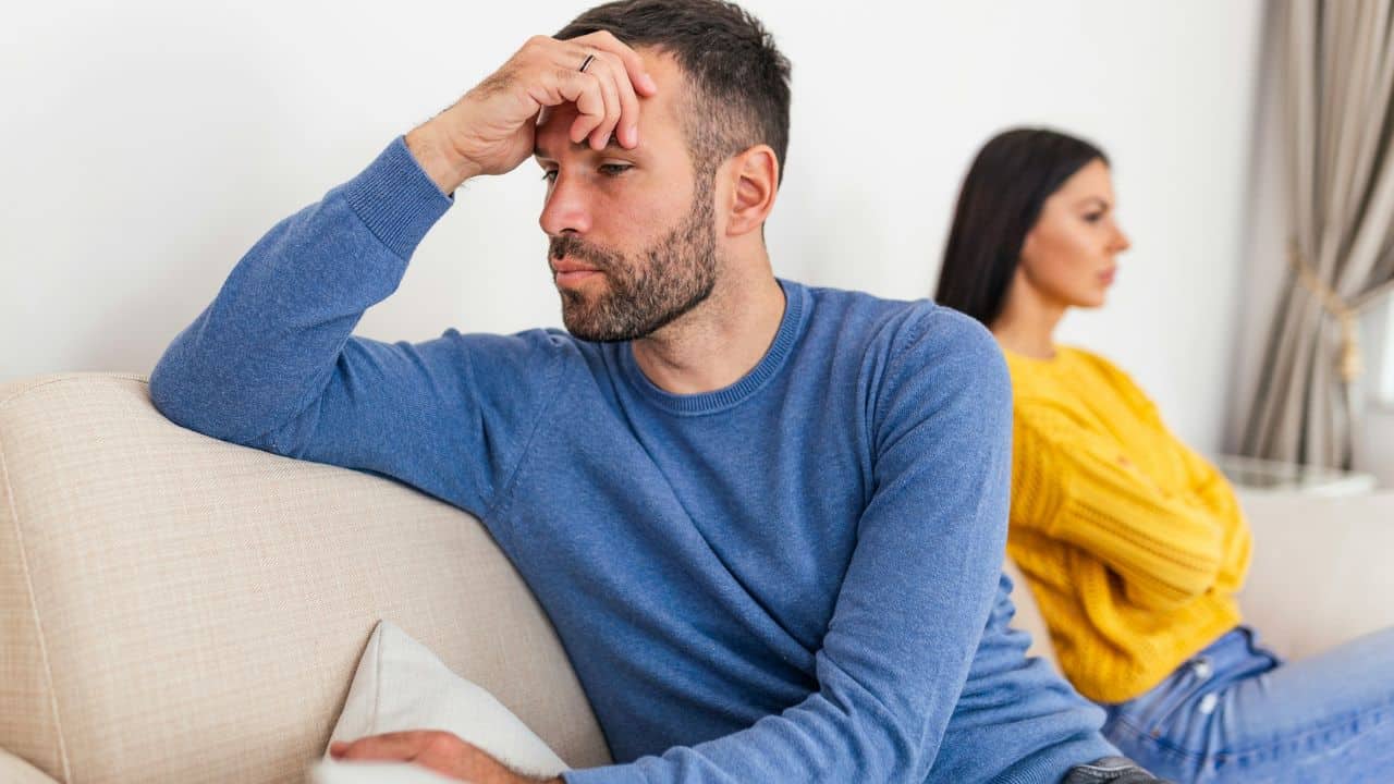 A man and woman sit on a sofa, looking away from each other with neutral expressions.
