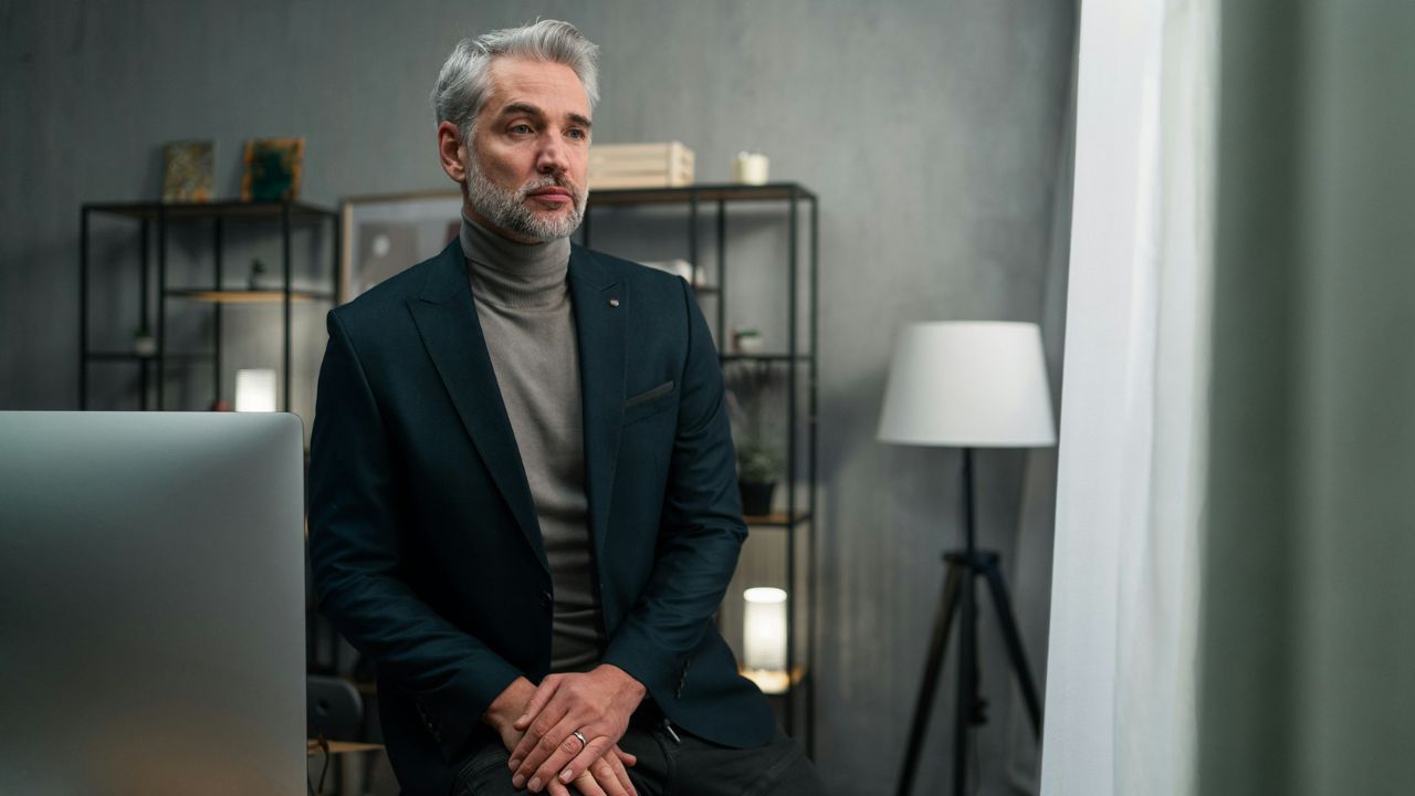 A handsome, gray-haired man in a turtleneck and suit jacket sitting in an office, looking pensive.