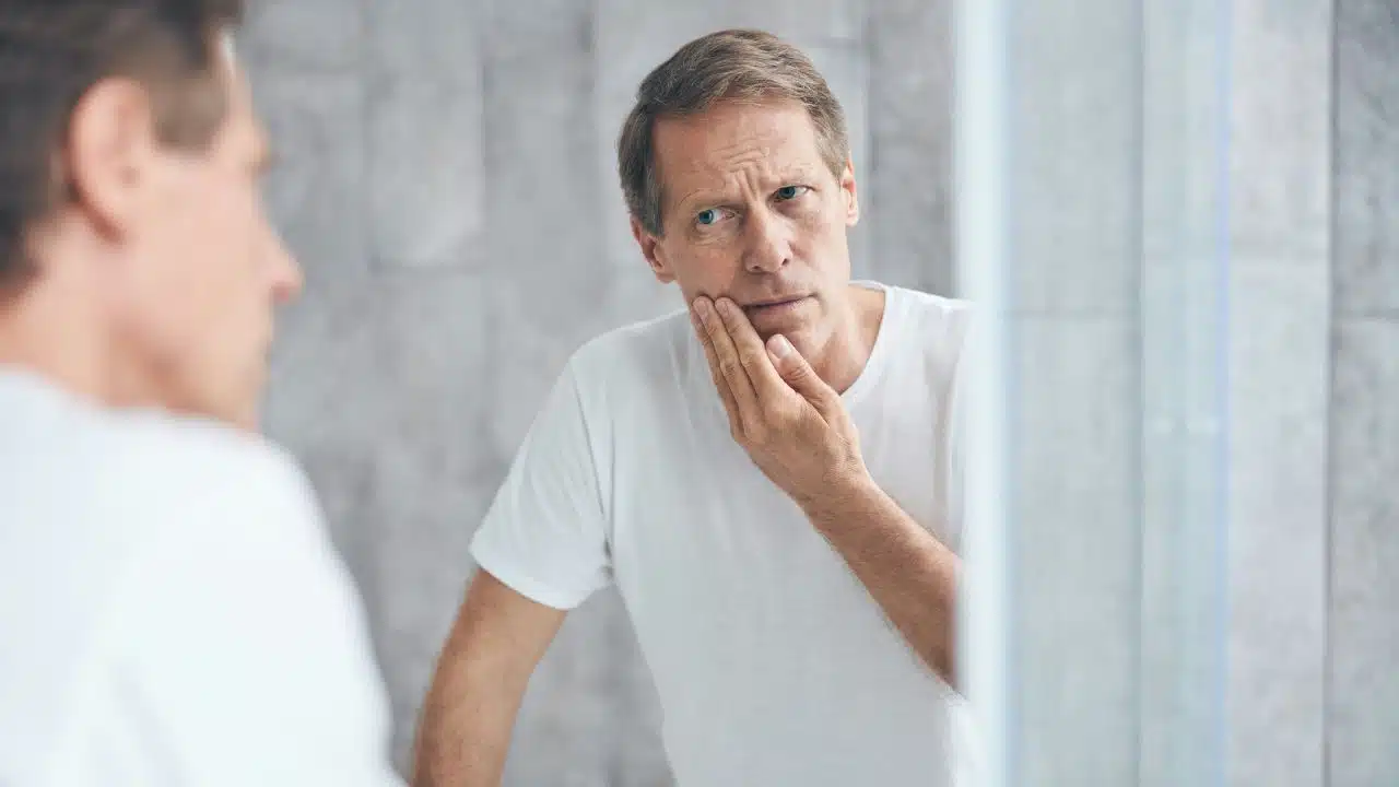 A man in a white shirt touches his face while looking into a bathroom mirror.