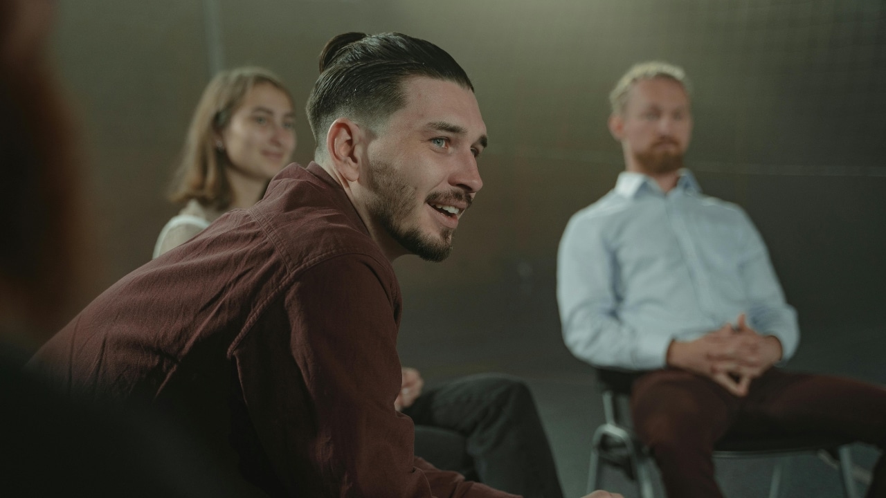 A man with a bun hairstyle speaks while sitting in a circle with others.