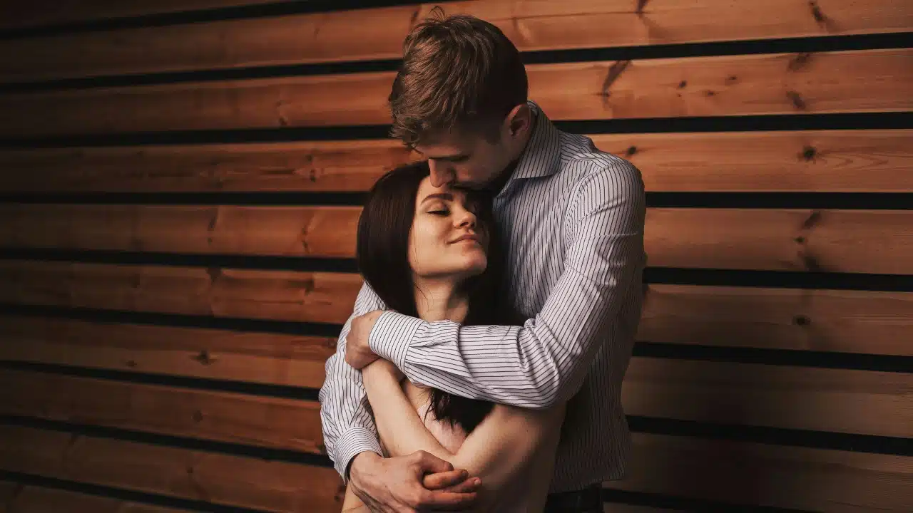 A man in a striped shirt embraces a woman from behind against a wooden wall.