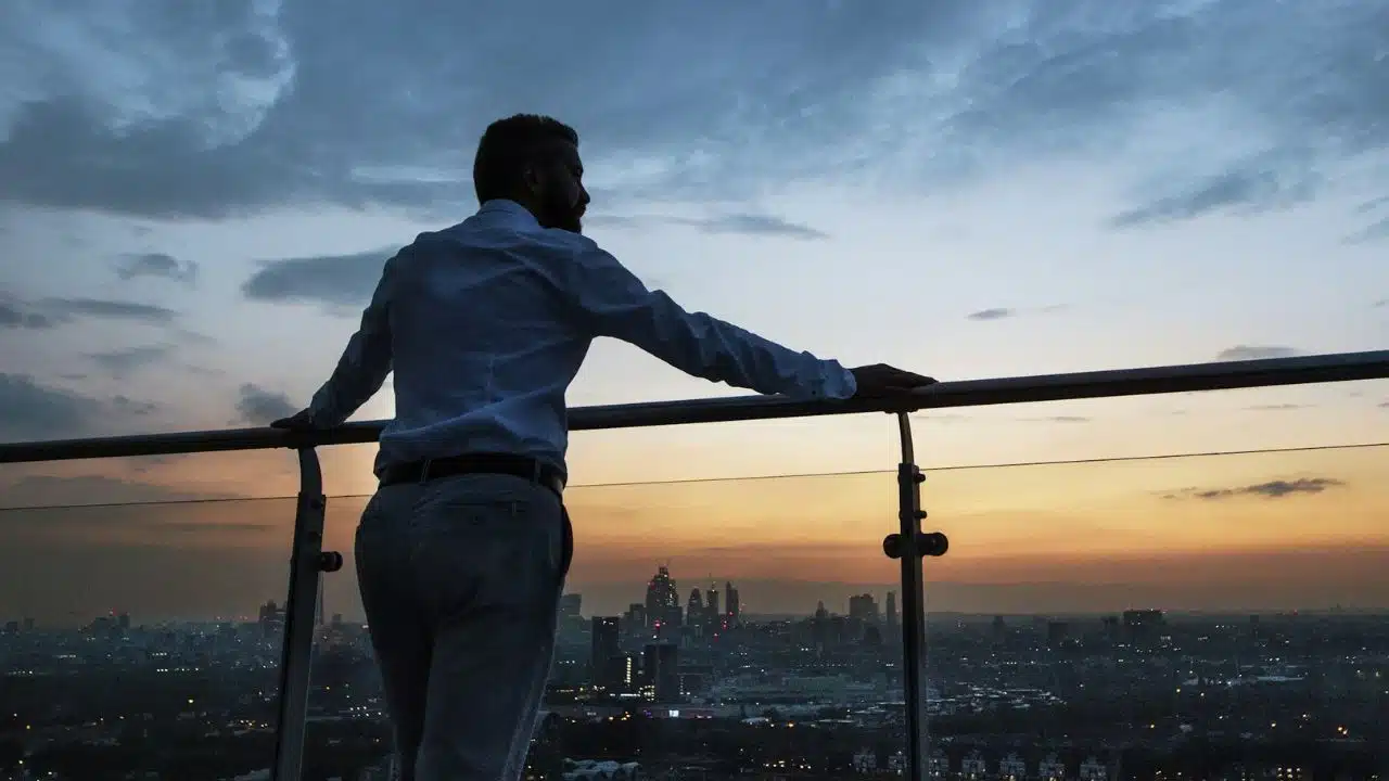 A man in a white shirt looks out over a city skyline during a sunset.