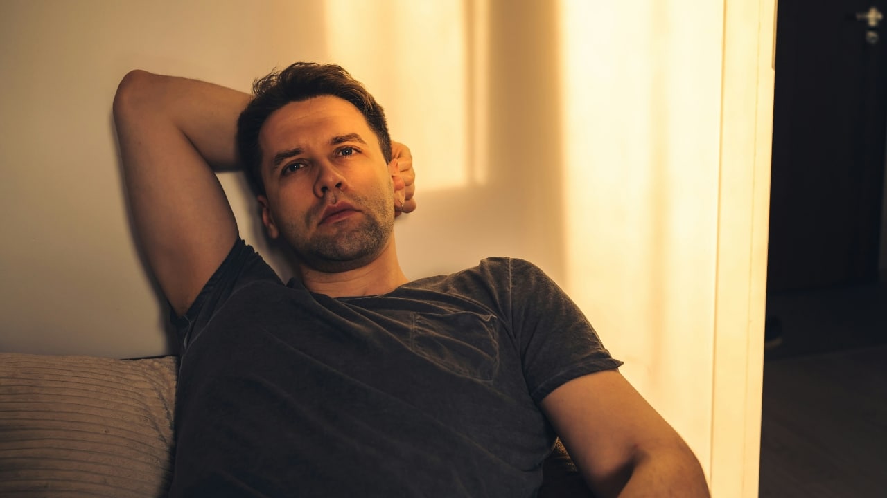 A man in a dark t-shirt leans against a wall with one arm raised.