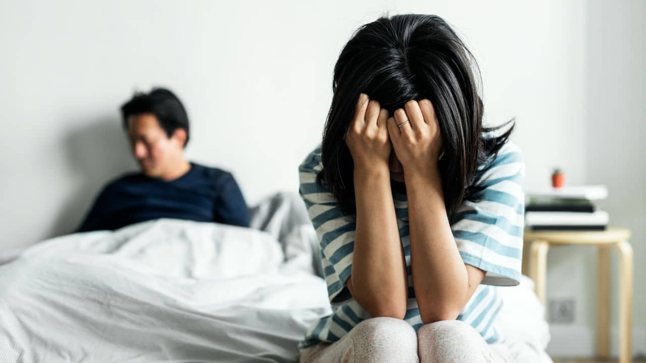 A person sits on a bed with hands over their face while another sits behind.