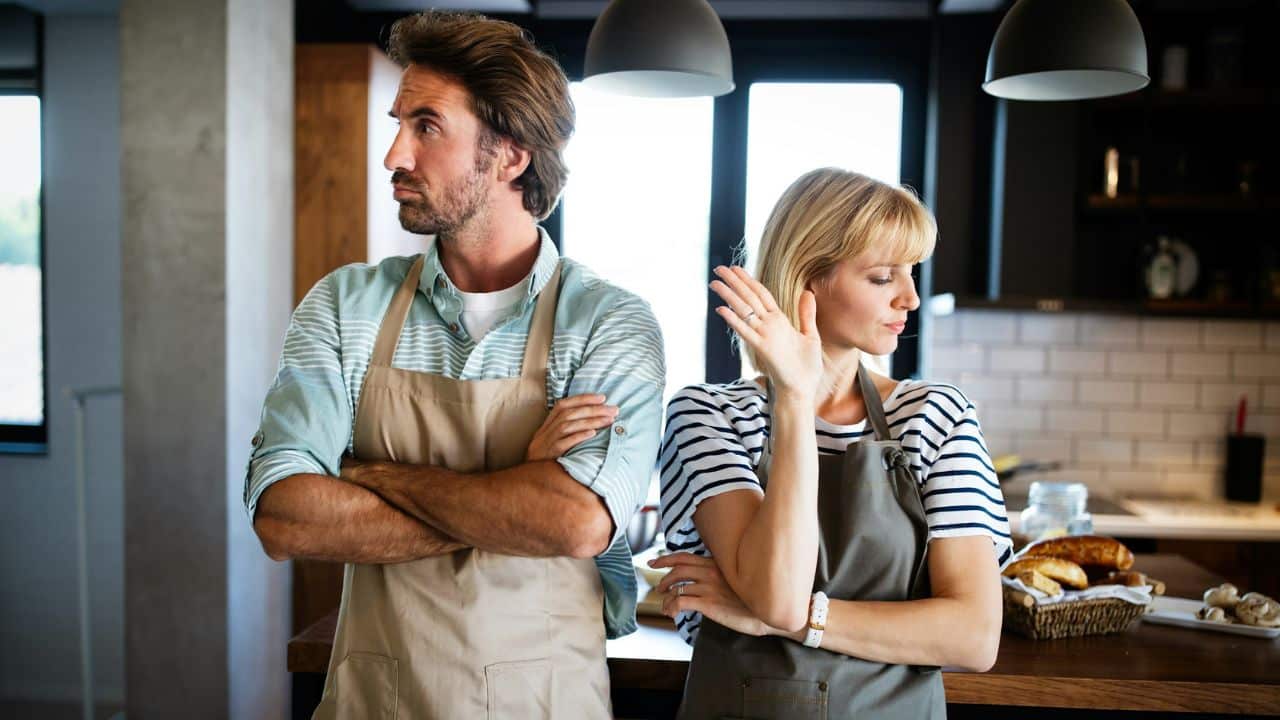 A frustrated couple wearing aprons stands back-to-back in a kitchen.