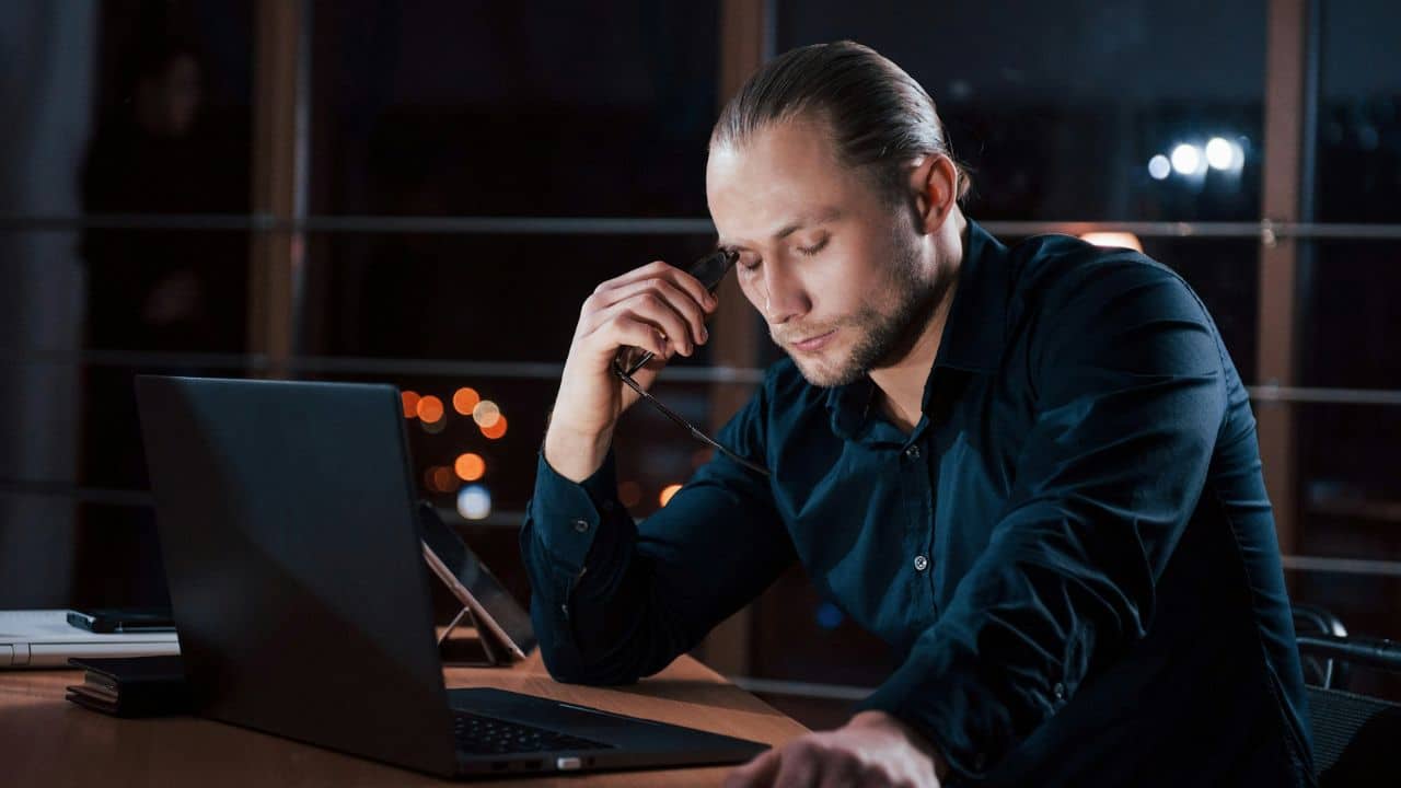 A man at a desk holds his glasses and touches his forehead near a laptop.