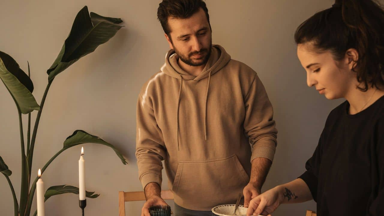A man in a tan hoodie looks down while a woman prepares food at a table.