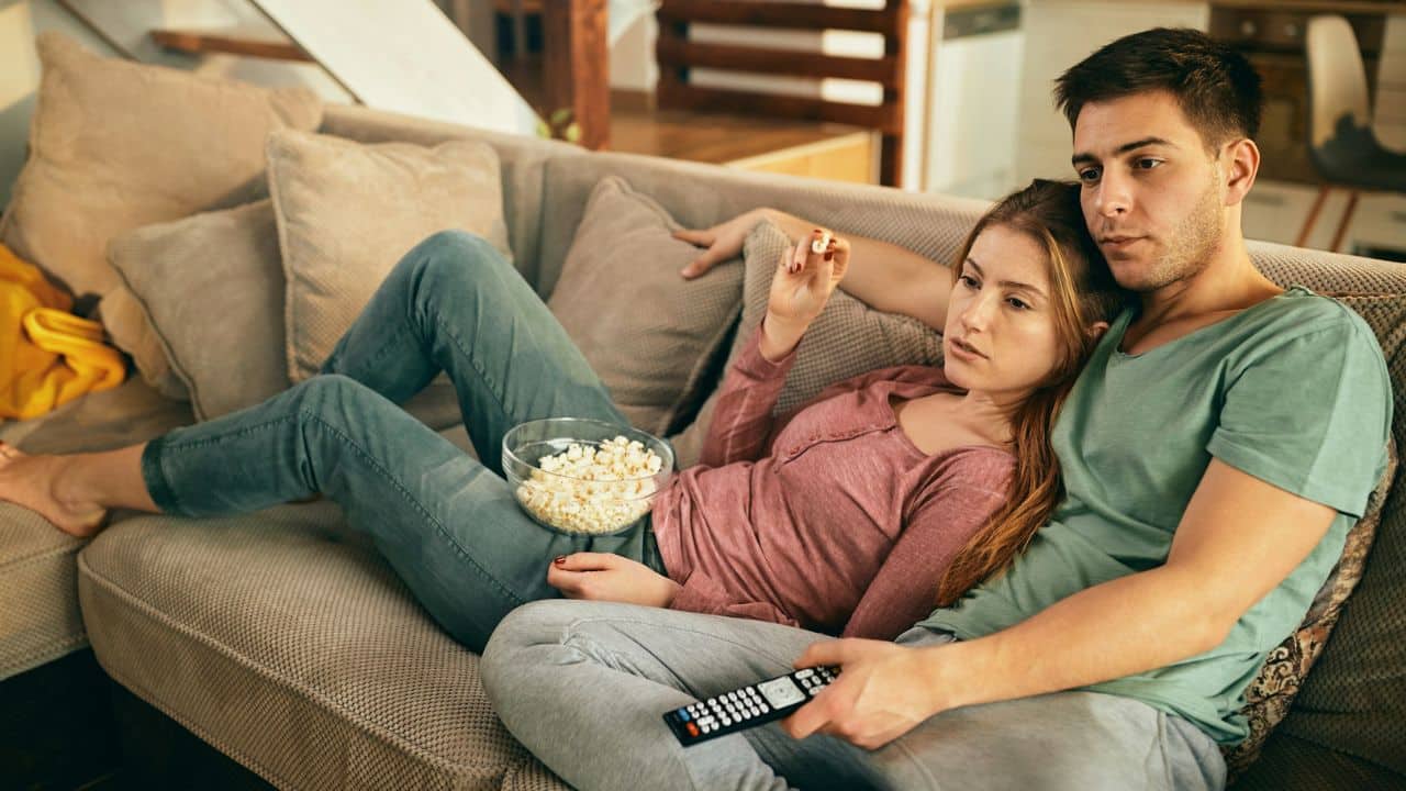 A man and woman sit on a sofa with popcorn and a remote control.