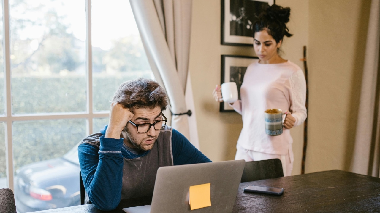 A man looks at a laptop while a woman stands behind him holding two mugs.