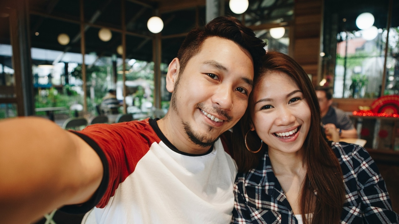 A smiling man and woman take a close-up selfie together in a brightly lit restaurant.
