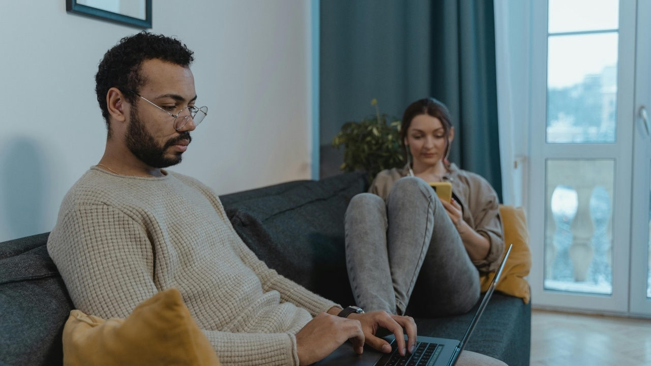 A man uses a laptop on a sofa while a woman sits behind him using a phone.
