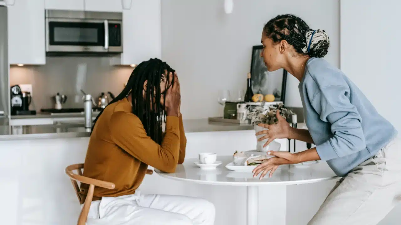 A woman gestures with her hands toward a man who is covering his face.