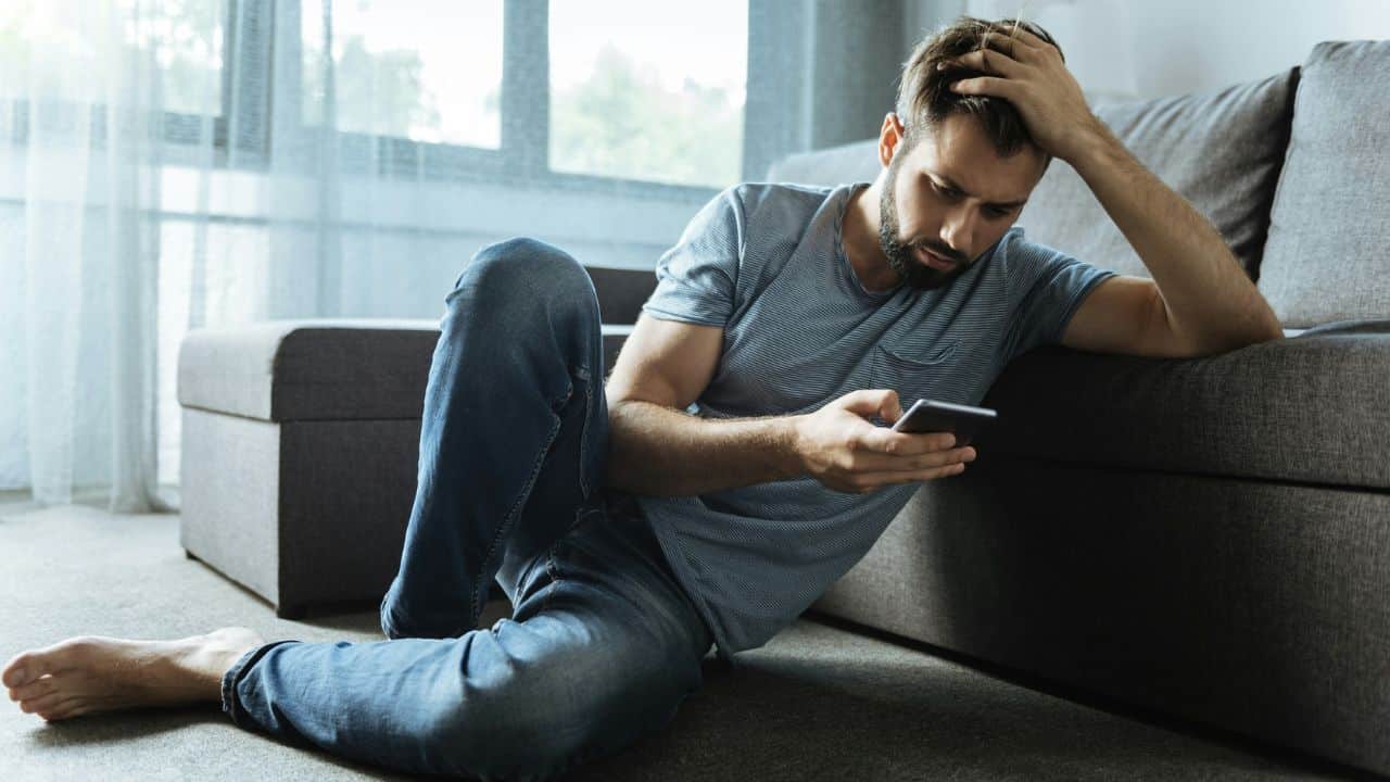 A man sits on the floor leaning against a sofa while looking at a phone.