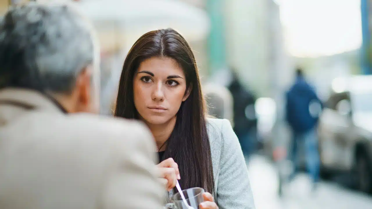 A woman with long dark hair sits outdoors, looking toward a man with grey hair.