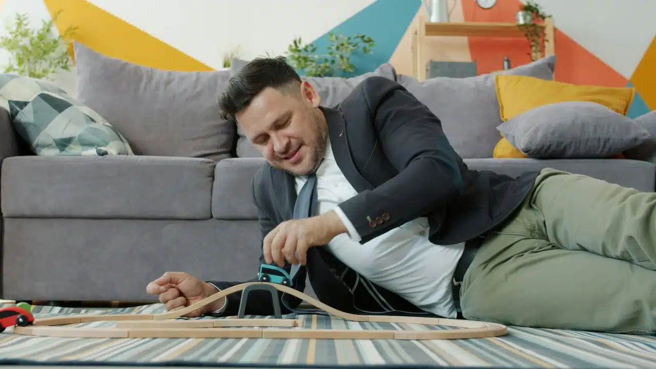A man in a suit lies on a rug and plays with a wooden toy train set.