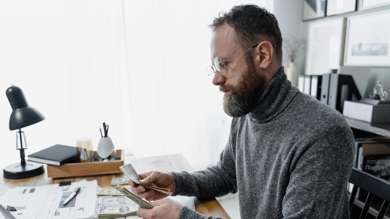 A bearded man in a gray turtleneck counts paper currency at a wooden desk.
