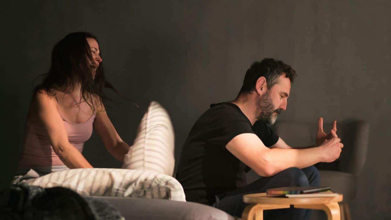 A woman throws a pillow while a man sits nearby with his head bowed down.