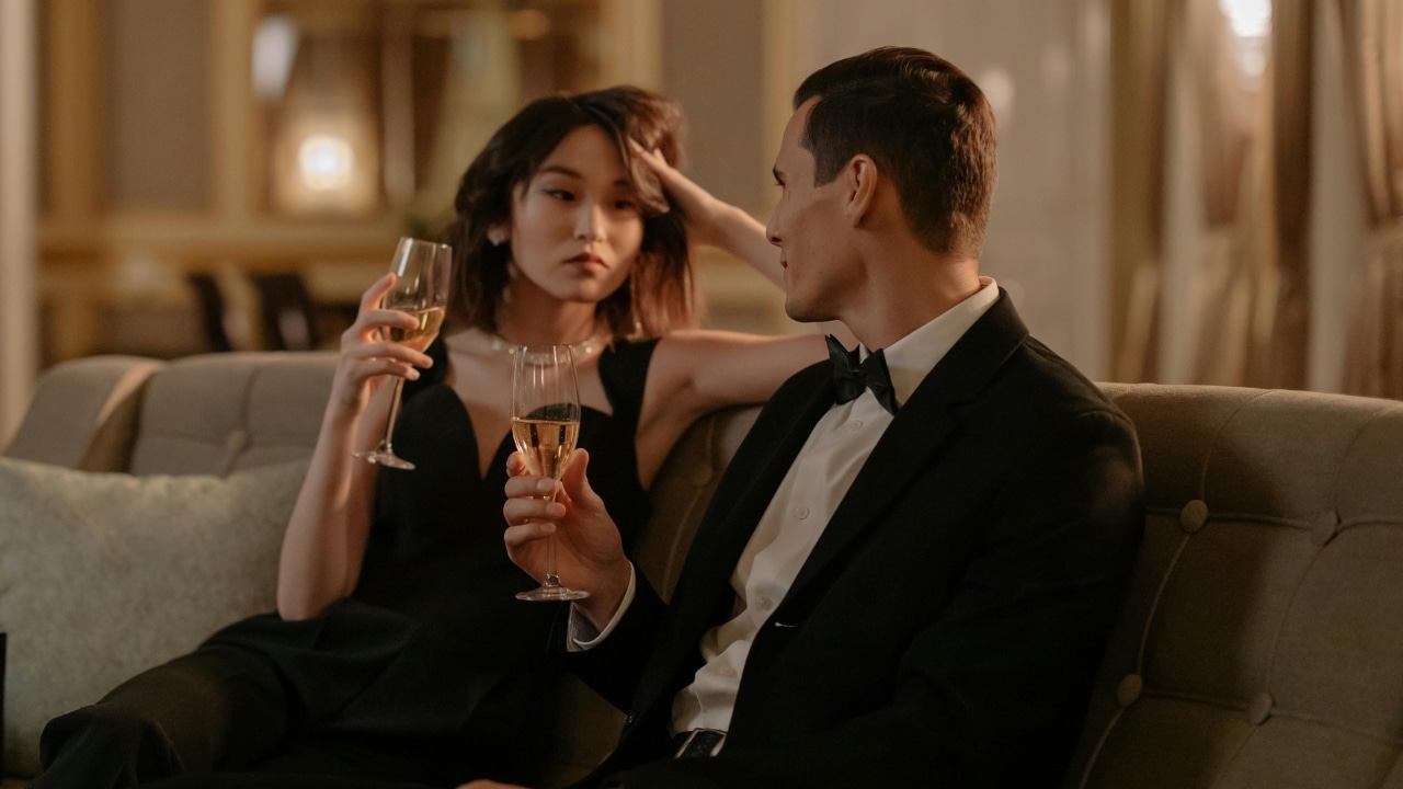 A man in a tuxedo and a woman in a black dress hold champagne glasses.