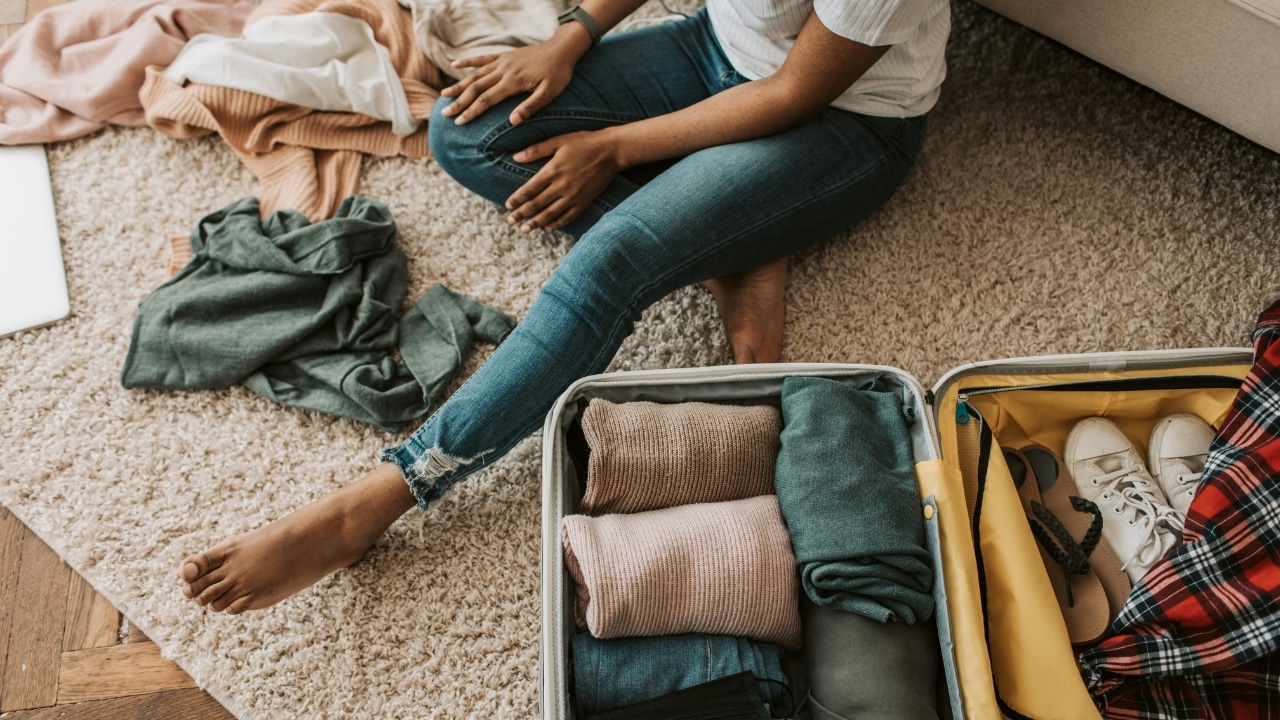 A person sits on a rug next to an open suitcase filled with folded clothes.