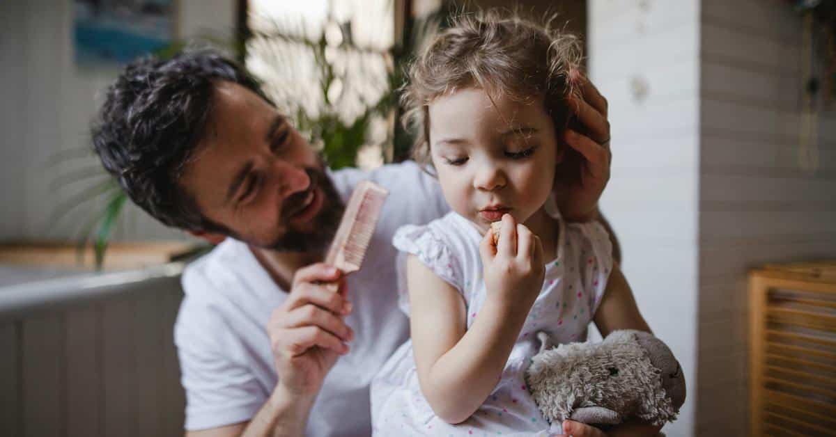 A father gently brushing a young child’s hair while she holds a stuffed toy.