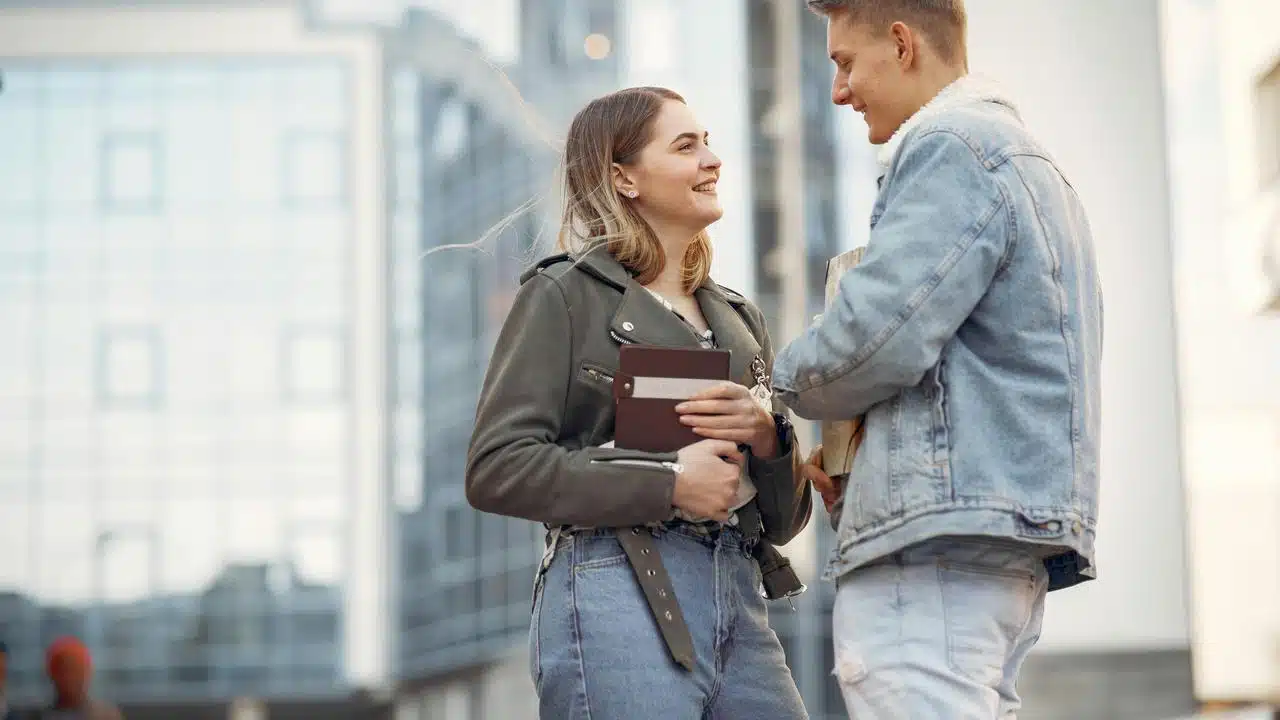 A young couple smiling and talking while holding books outdoors.
