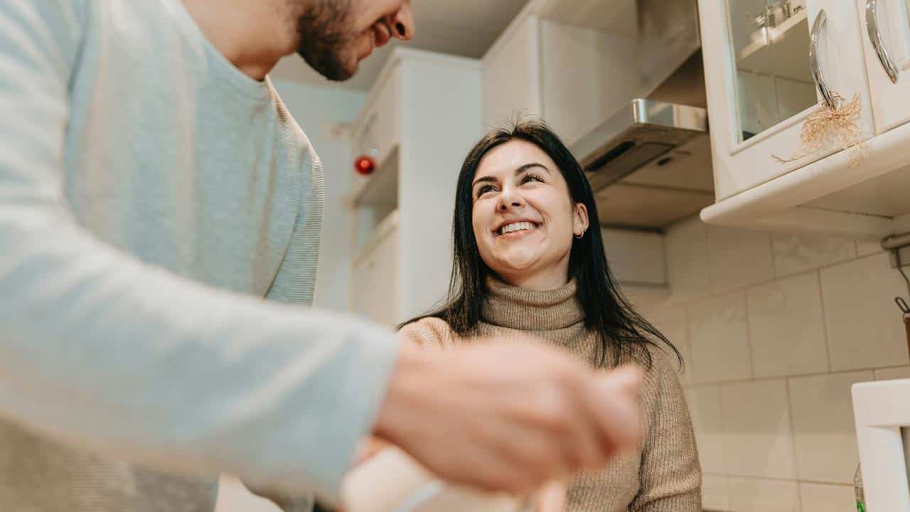 A couple smiles and talks while cooking together in a kitchen.