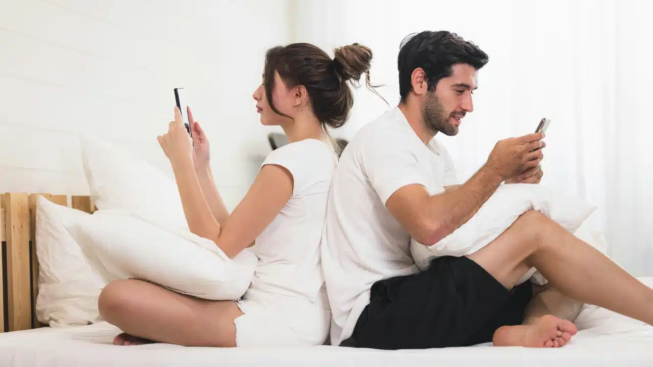 A man and woman sit back-to-back on a bed, each looking at their own smartphone.