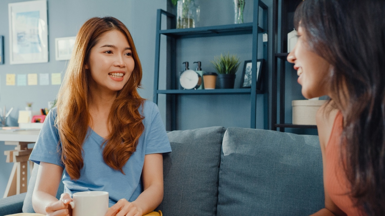 Two women sitting on a couch indoors, with one woman smiling and holding a mug.
