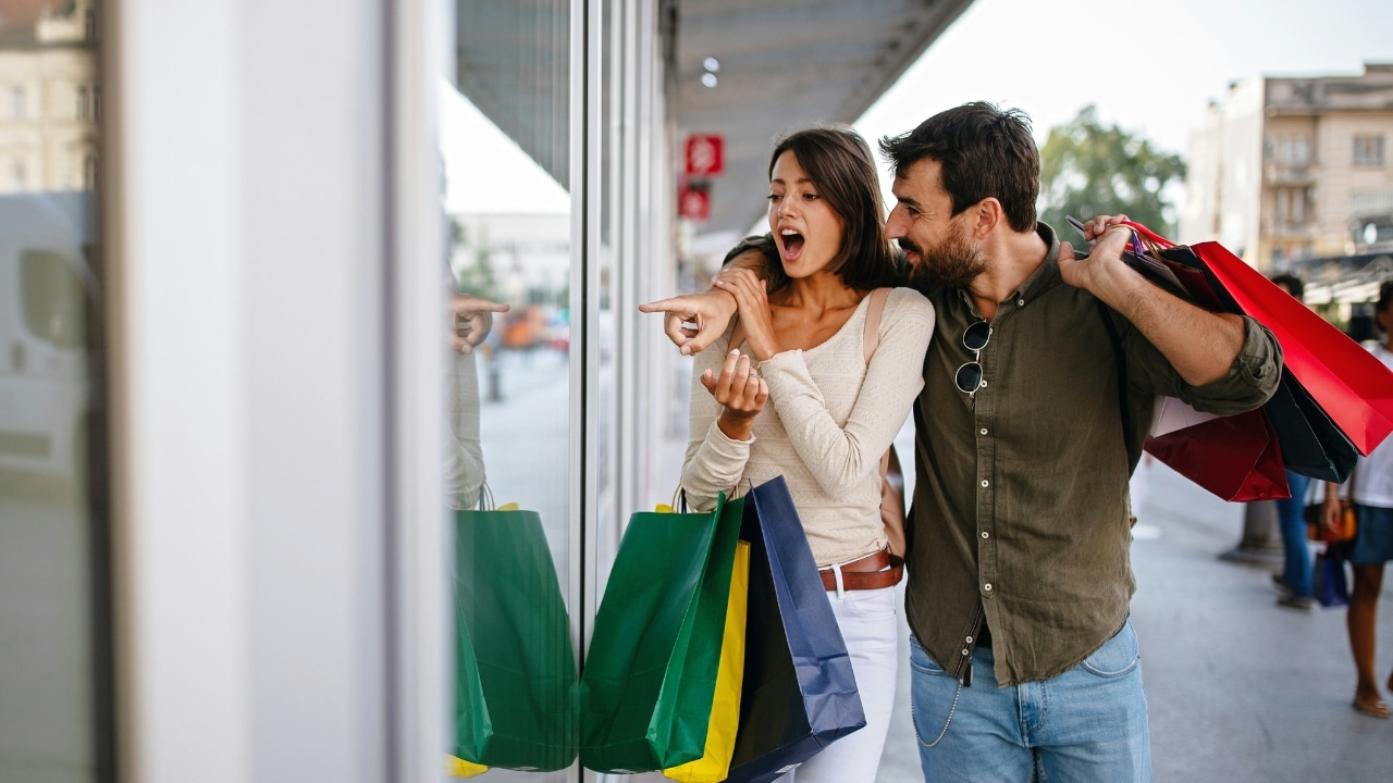 A man and woman walk outdoors carrying several colorful shopping bags while looking at a window.