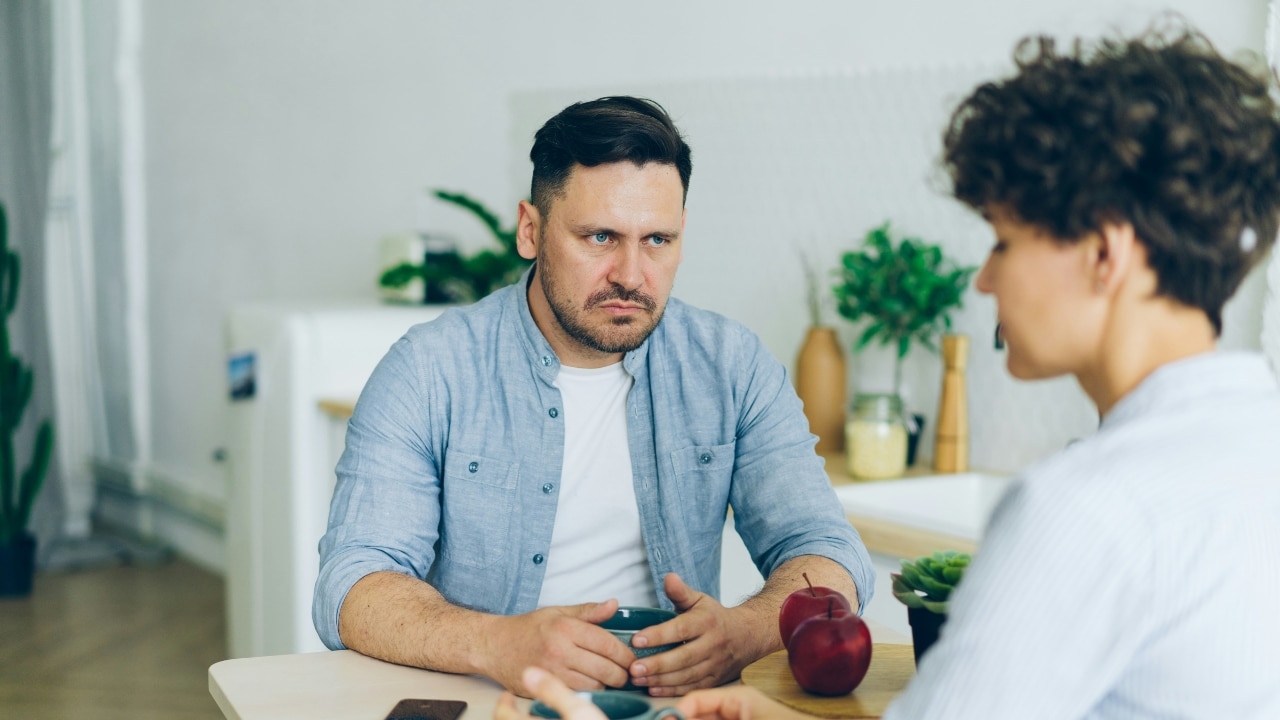 A man with a frown looks at a woman across a table with two red apples.