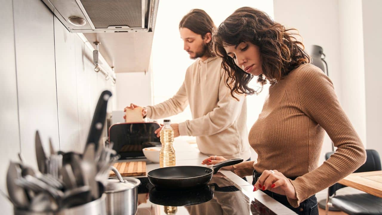 A woman stands at a stove while a man prepares toast in a kitchen nearby.