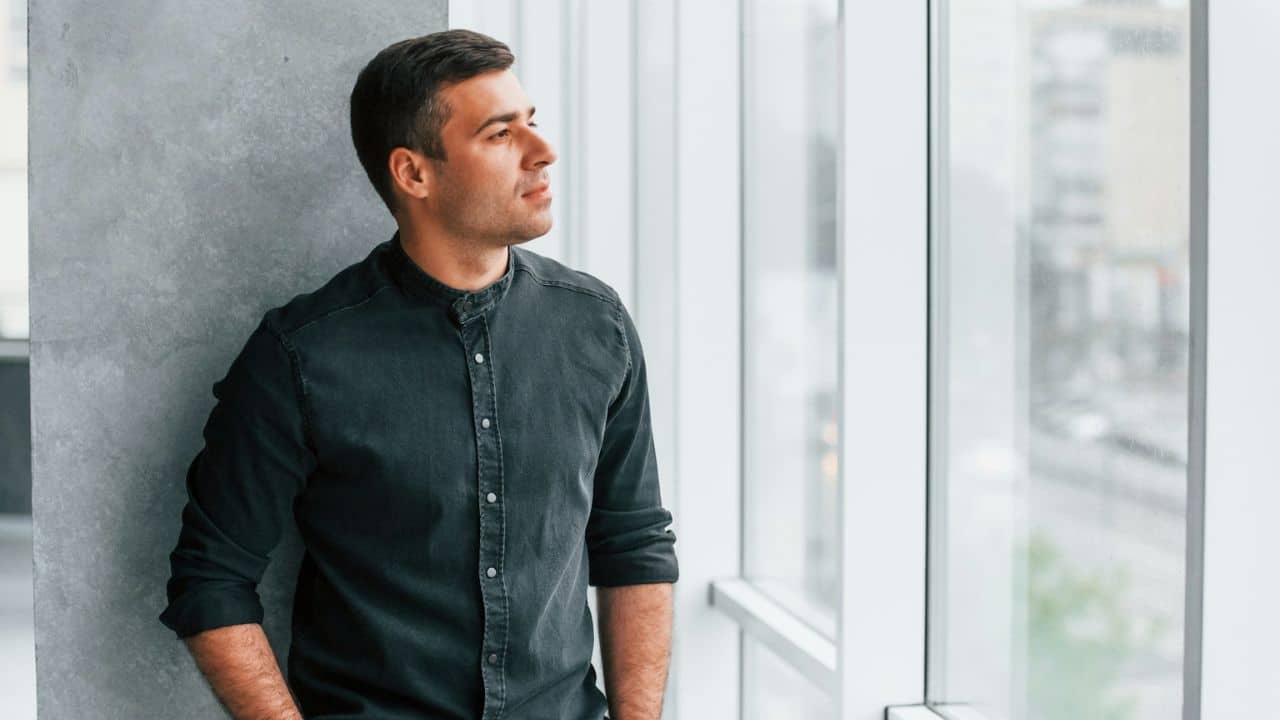 A man in a dark shirt stands by a window, looking out thoughtfully.