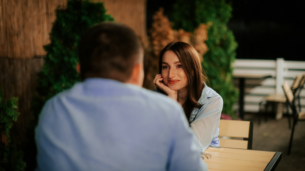 A woman smiling and looking at a man across a table on an outdoor patio at night.