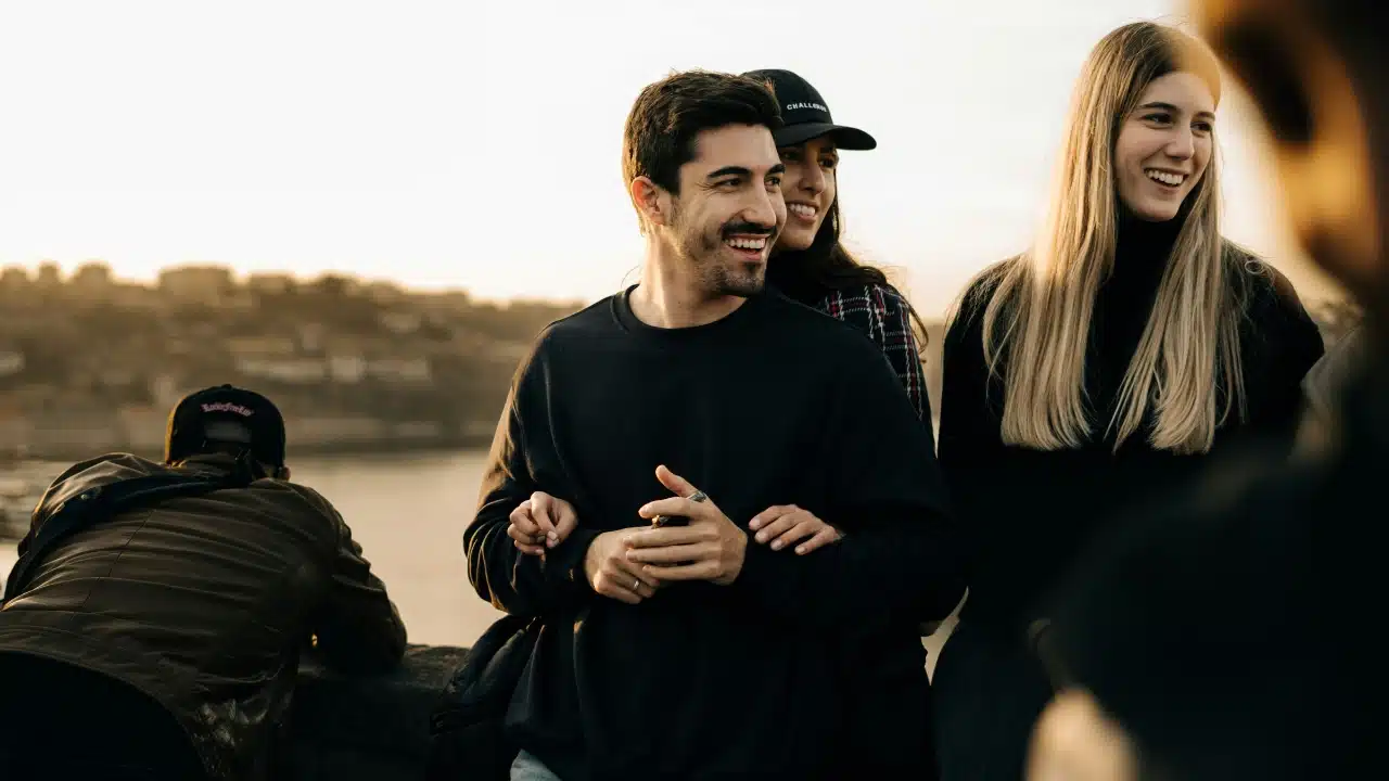 A smiling man and woman stand together outdoors with a scenic city background at sunset.