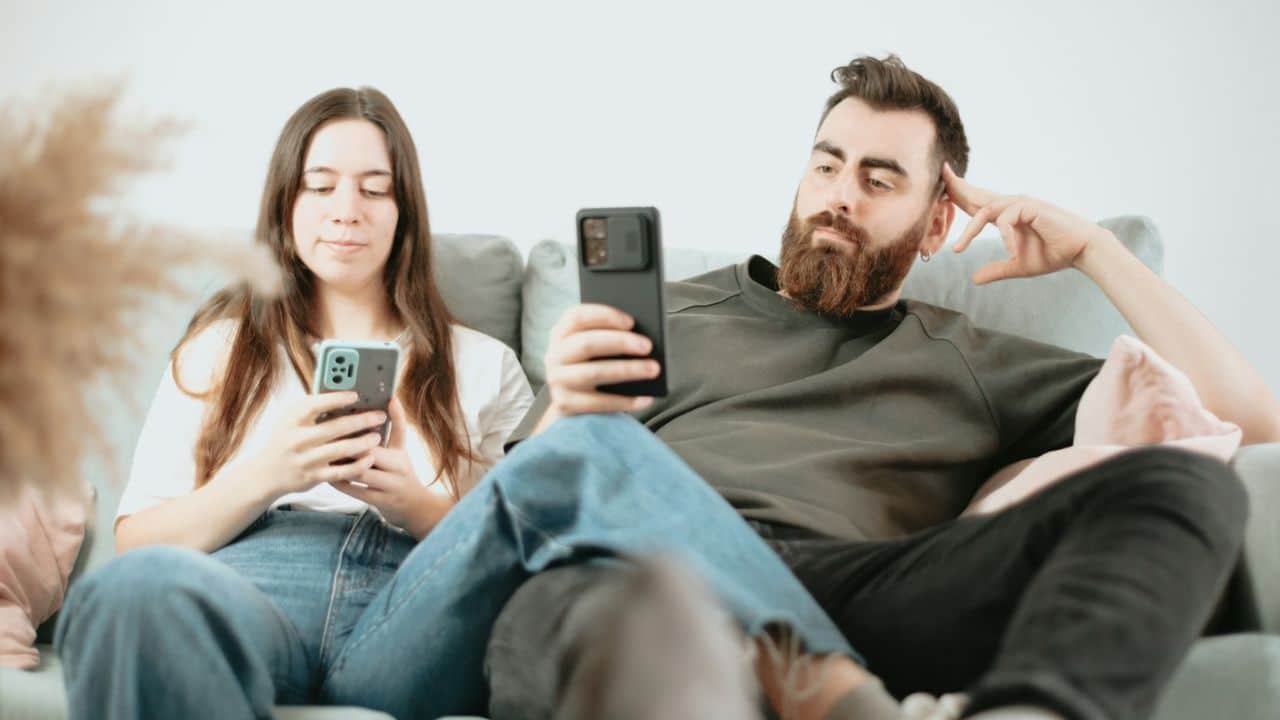 A man and woman sit on a couch, each looking at their own smartphone screen.