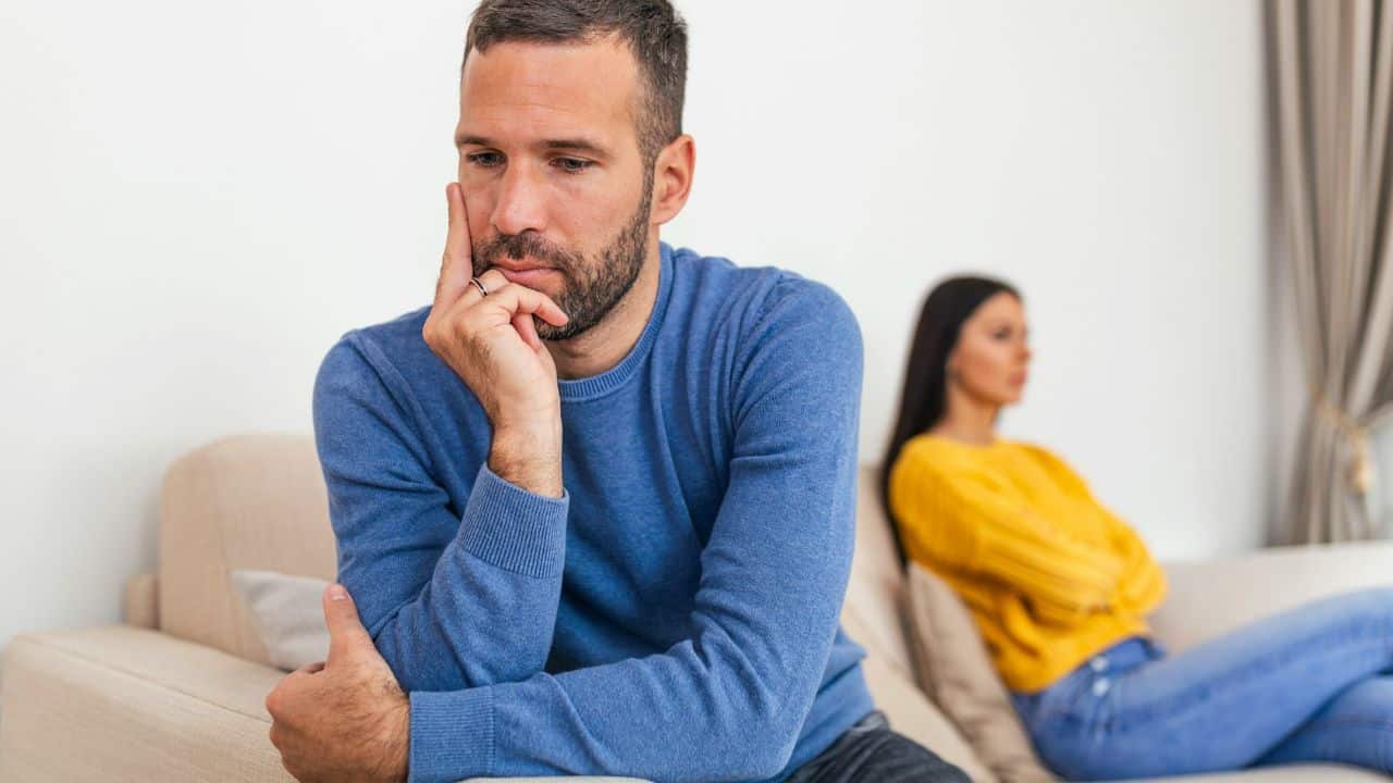 A man sitting on a sofa looking down while a woman sits blurred in the background.